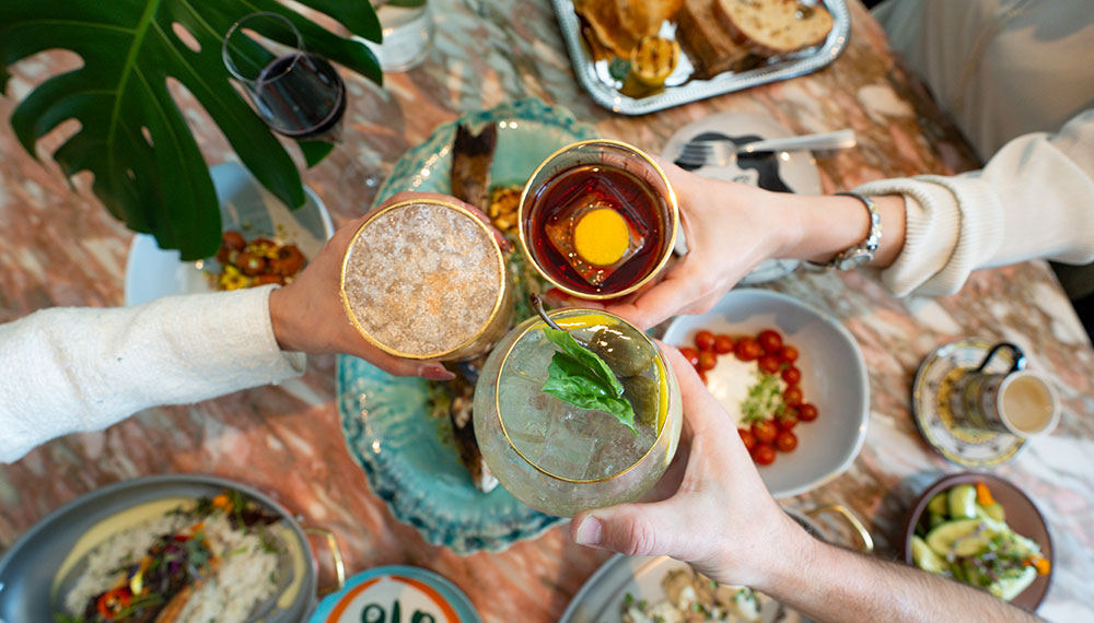 3 hands cheersing with cocktail over a selection of food items